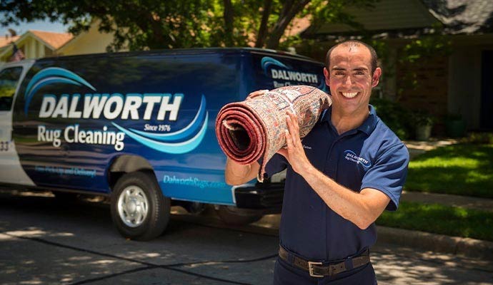 A smiling Dalworth Rug Cleaning professional carrying a rolled oriental rug in front of a company service van