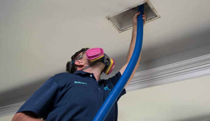A professional technician wearing a respirator mask while using a high-powered blue vacuum hose to clean a ceiling air vent