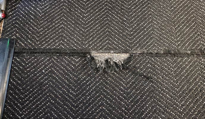 A top-down view of a dark herringbone patterned carpet showing a frayed and separating seam in the center