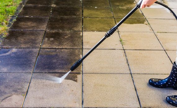 Person using a pressure washer to clean dirty concrete tiles outdoors.